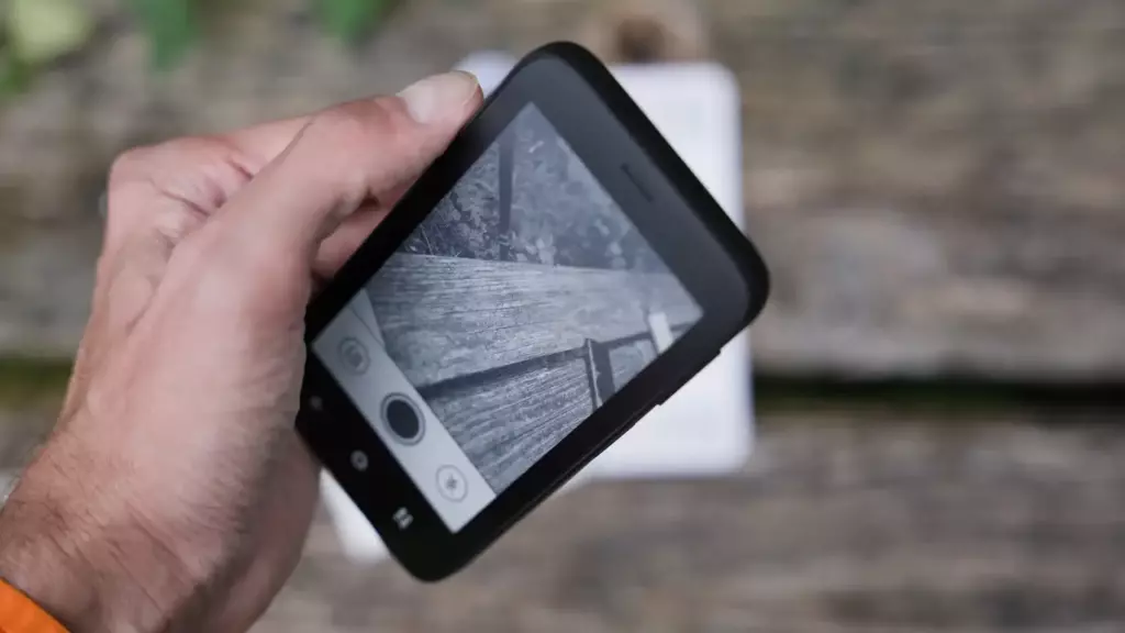 Смартфон Mudita Kompakt в руке, на E-Ink экране открыт интерфейс камеры. Пользователь снимает деревянную поверхность на фоне природы. Видны кнопки управления съёмкой.